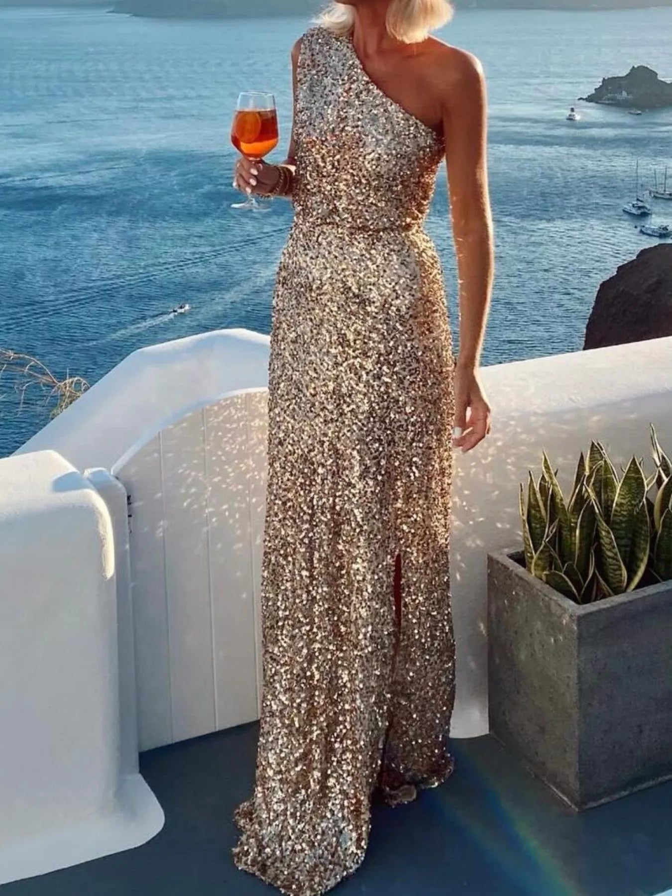 Model wearing a sequin one-shoulder maxi dress holding a drink by the sea, showcasing evening elegance.