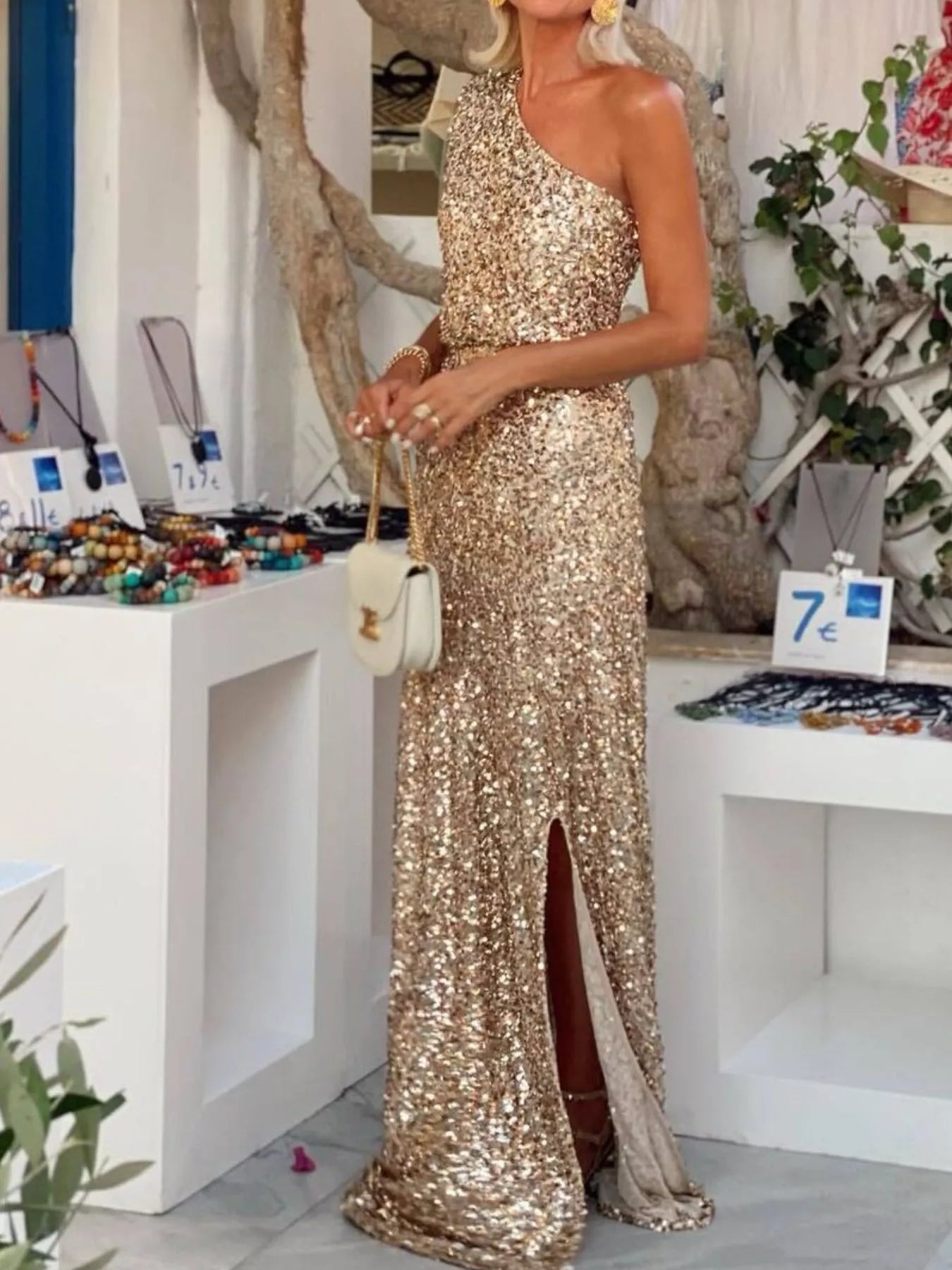 Elegant woman wearing a sequin one-shoulder maxi dress with a side slit in a chic boutique setting.